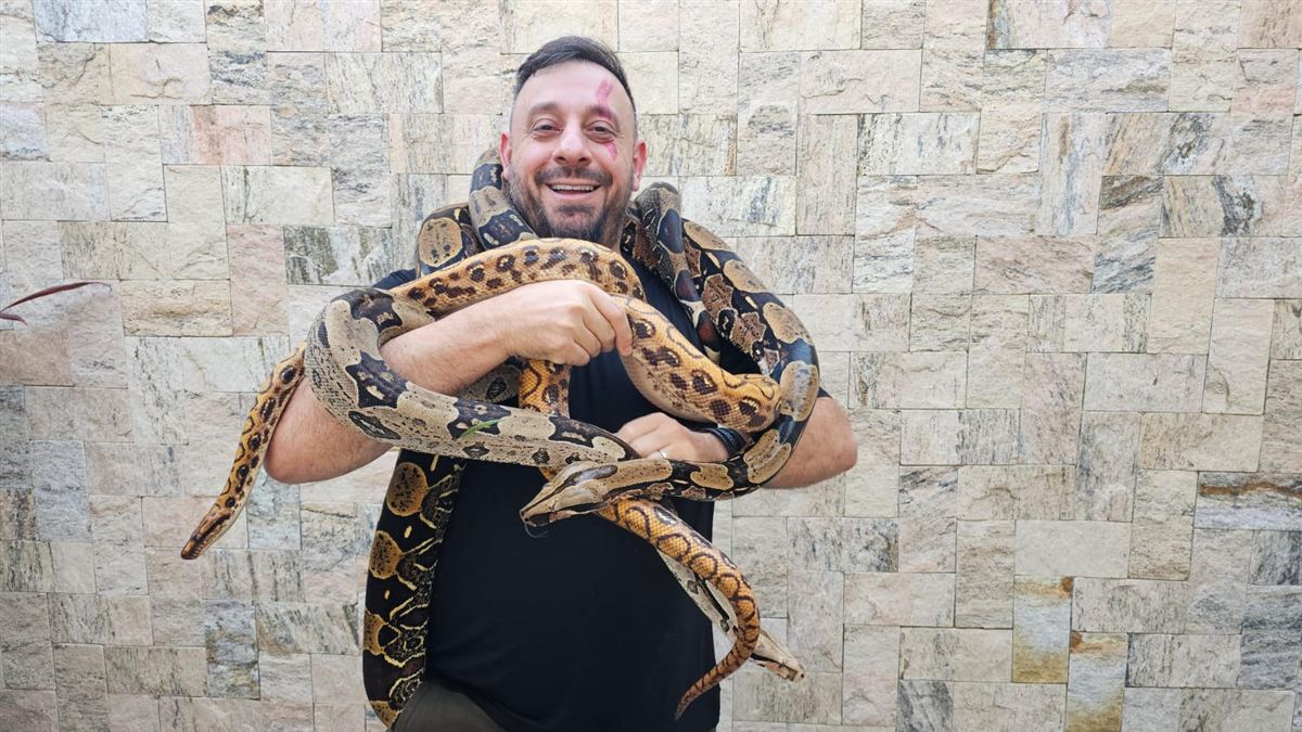 Biólogo Henrique com uma jiboia enrolada nos braços e no pescoço, sorrindo para a câmera